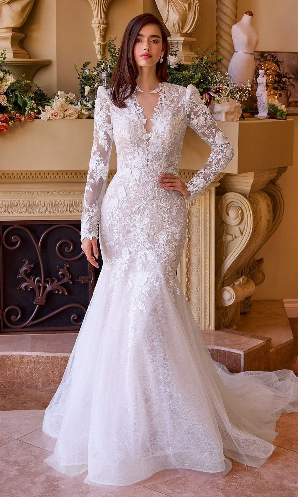 A woman stands indoors wearing the Andrea and Leo WL036 long wedding dress, a fitted white lace mermaid gown with floral details and an elegant, dramatic silhouette.